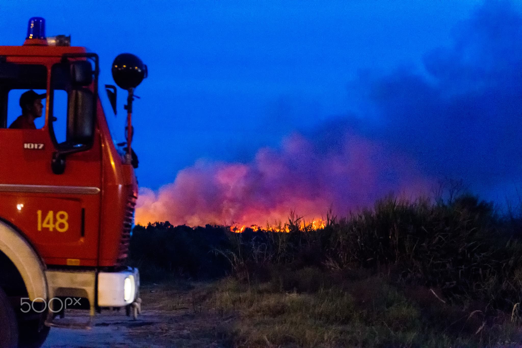 Bombero en camión de emergencia observa un incendio forestal activo al atardecer con llamas y humo visible en el horizonte.