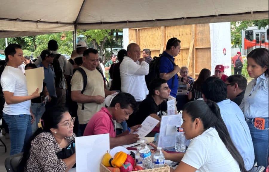 Personas bajo una carpa participan en una feria de empleo donde interactúan con reclutadores en mesas de atención.