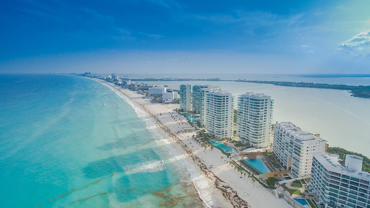 Vista aérea de la zona hotelera de Cancún con playas turquesas, edificios de alta densidad y el mar de fondo bajo cielo despejado.