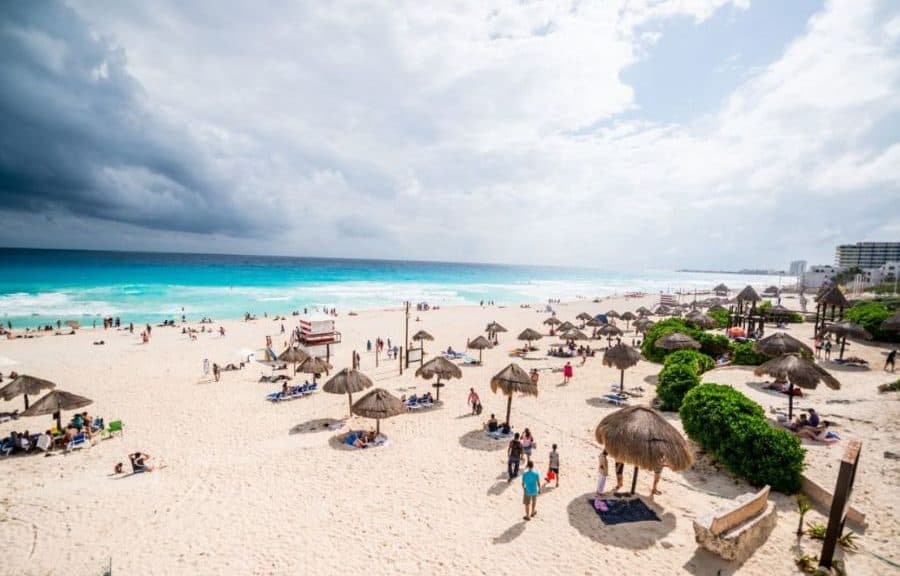Playa concurrida de Cancún con arena blanca, sombrillas de palapa, turistas y mar turqueso bajo cielo nublado.