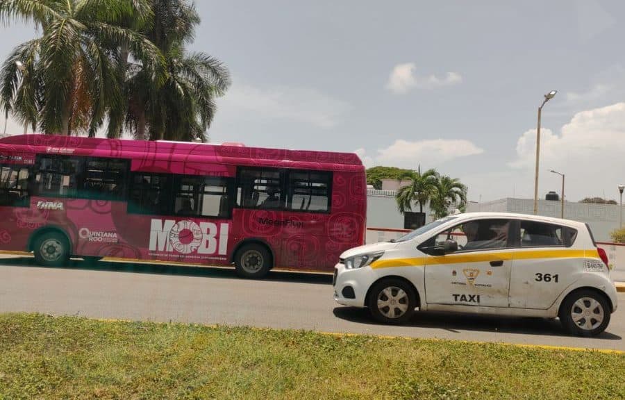 Taxi blanco con número 361 estacionado junto a un autobús de transporte público de color magenta en una calle de Chetumal.
