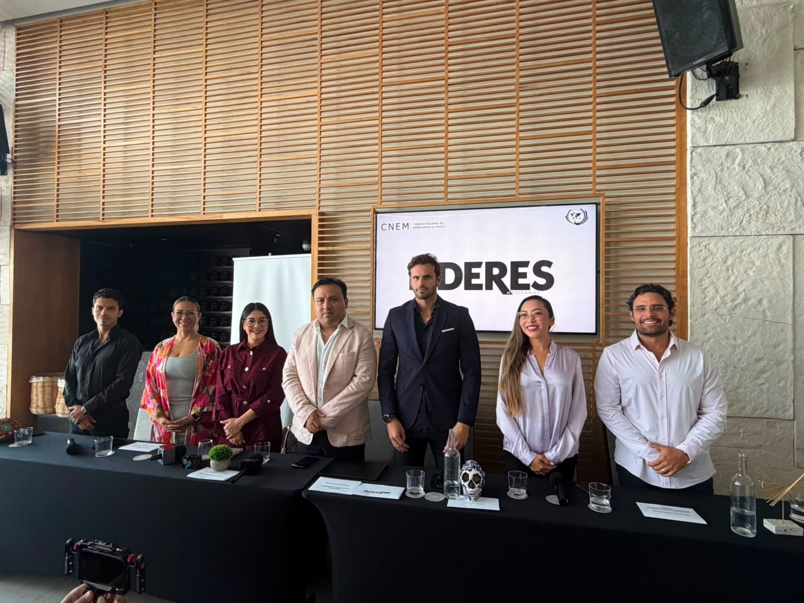 Siete personas en traje de negocios de pie frente a una mesa con micrófono durante una conferencia, con pantalla de fondo que muestra texto sobre emprendimiento.