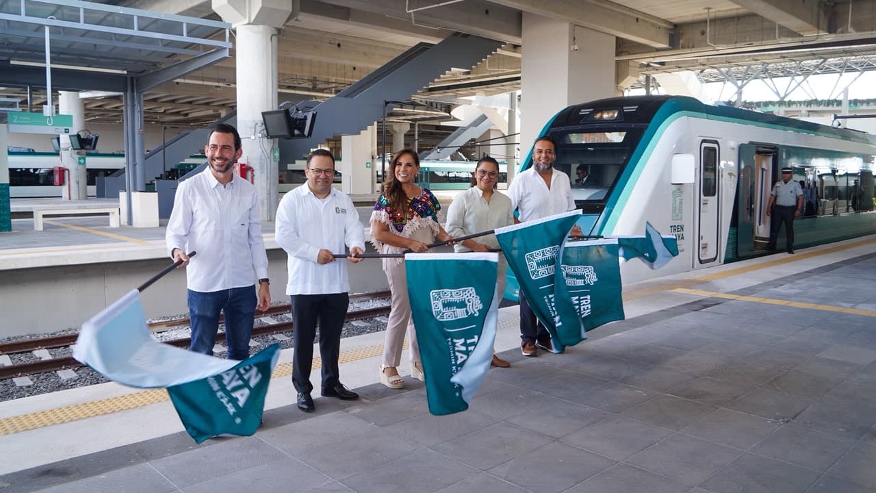 Cinco personas en traje formal sostienen una cinta durante la inauguración de un tren moderno de color turquesa en una estación.