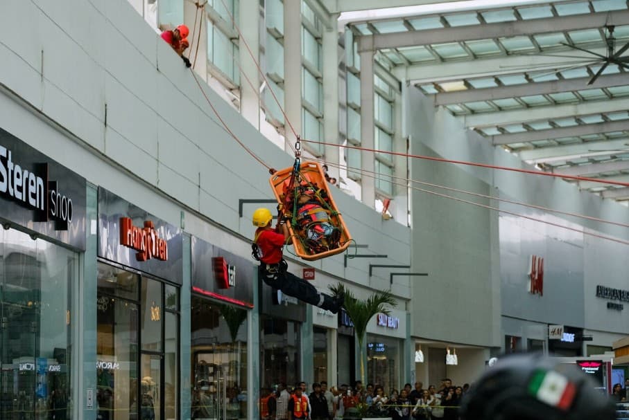Simulacro de rescate en altura dentro de un centro comercial con personal en equipo de seguridad bajando a una persona en camilla naranja mediante cuerdas.