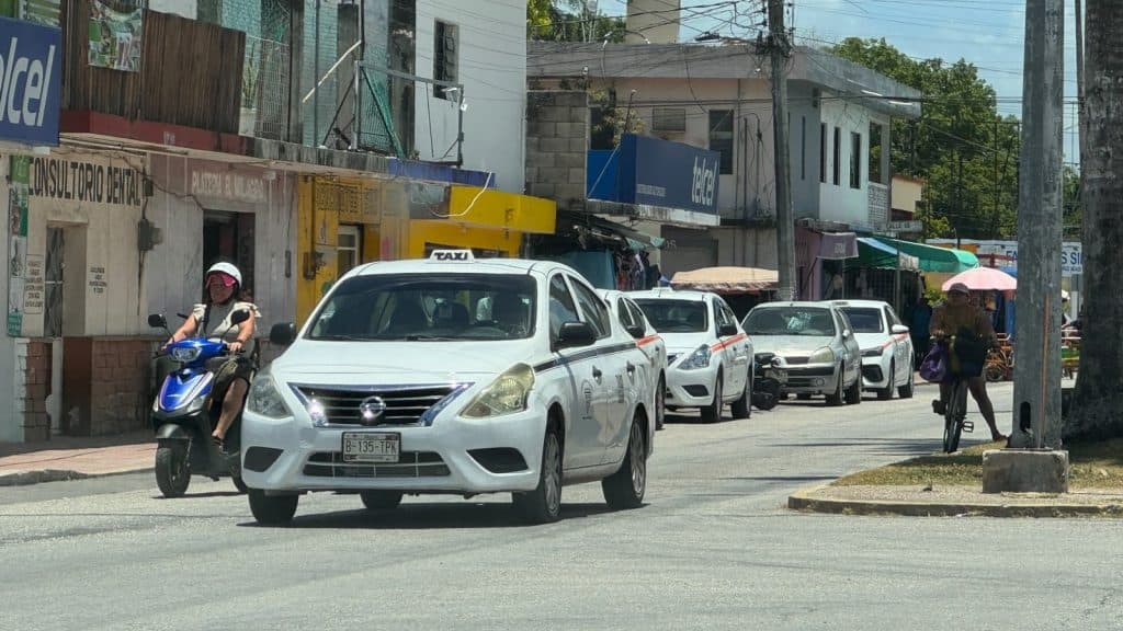 Taxis blancos de radio taxi estacionados en una calle de Felipe Carrillo Puerto con comercios locales de fondo.