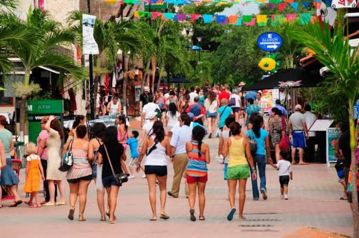 Calle peatonal concurrida en Playa del Carmen con turistas caminando, tiendas y decoraciones festivas de colores.