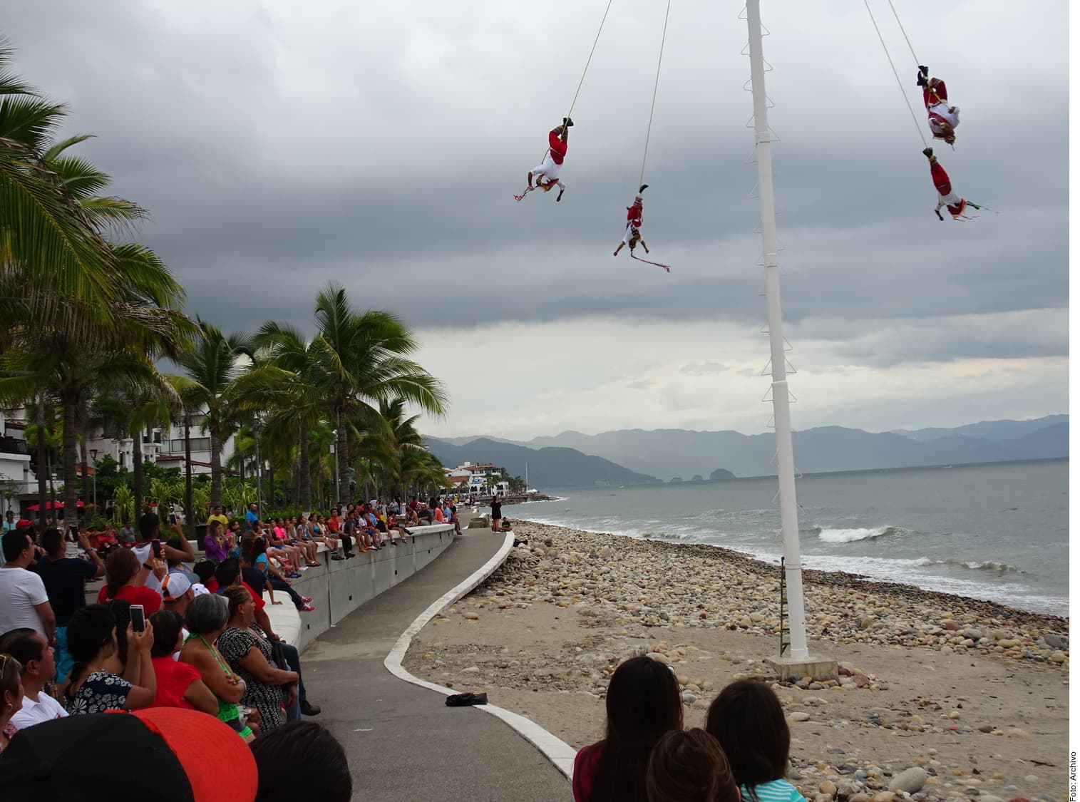 Personas realizan rappel desde un poste en una playa mexicana mientras una multitud observa desde la orilla, con palmeras y montañas de fondo.