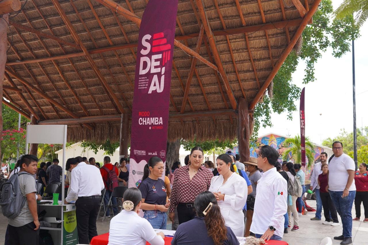 Personas asisten a una feria de empleo bajo estructura de palapa en Playa del Carmen, con stands informativos y personal disponible.