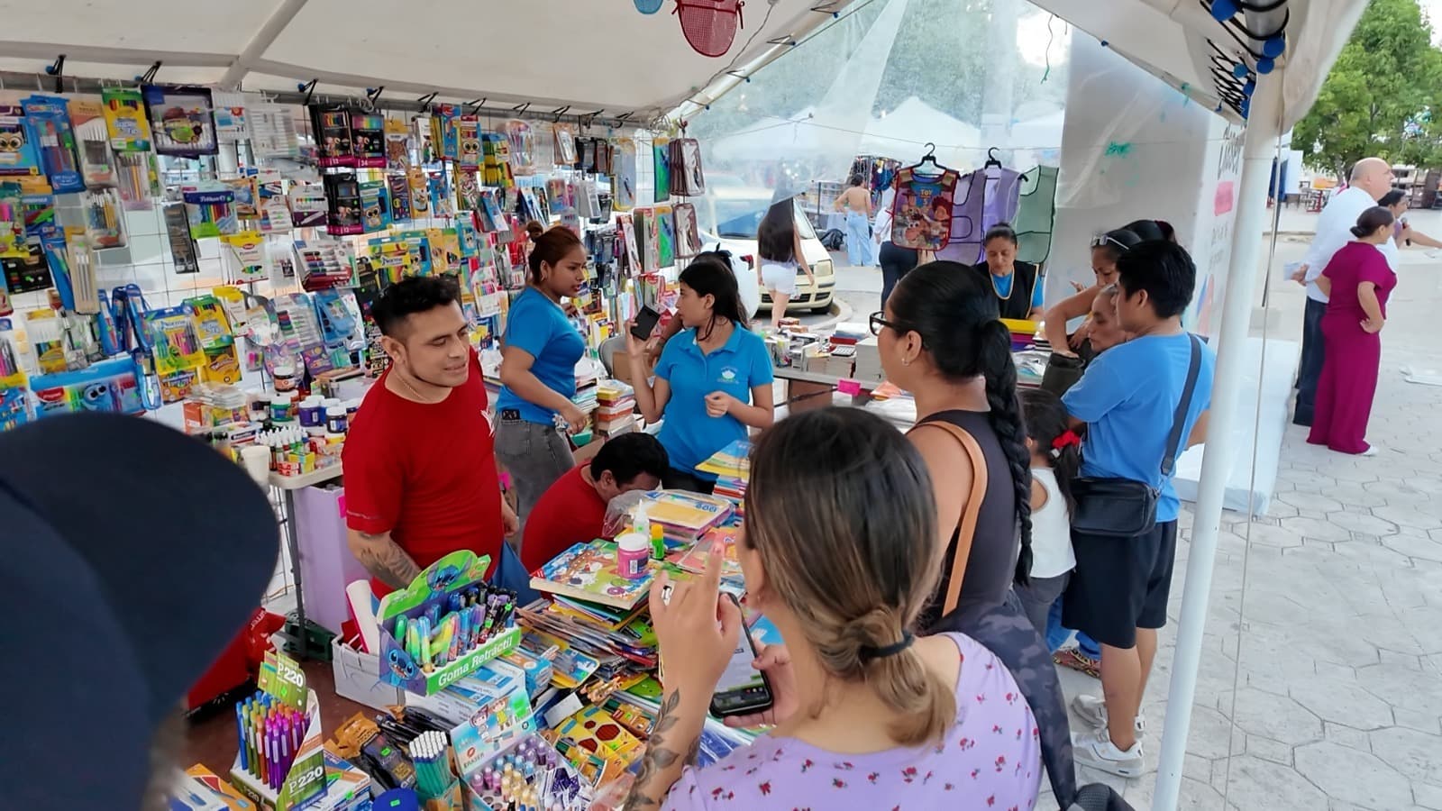 Personas compran útiles escolares y libros en un puesto comercial bajo carpa en una zona exterior.