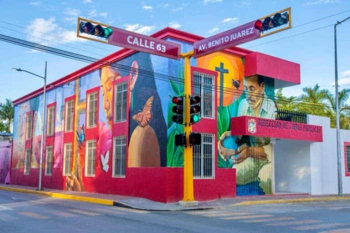 Edificio público pintado con murales coloridos ubicado en la esquina de Calle 63 y Avenida Benito Juárez, con señalización vial visible.