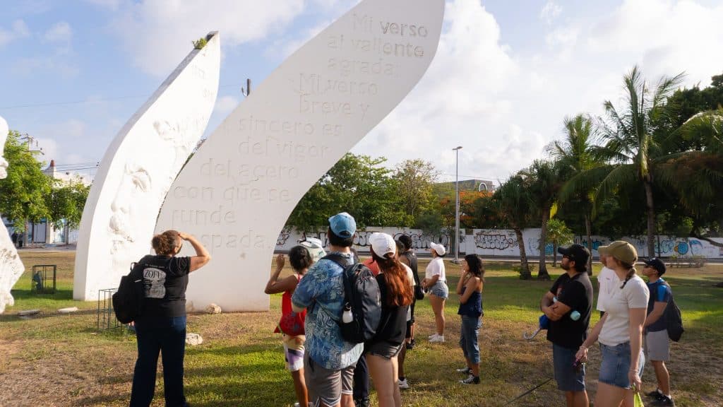 Grupo de turistas observa una escultura monumental blanca con texto grabado en la Zona Fundacional de Cancún bajo cielo nublado.