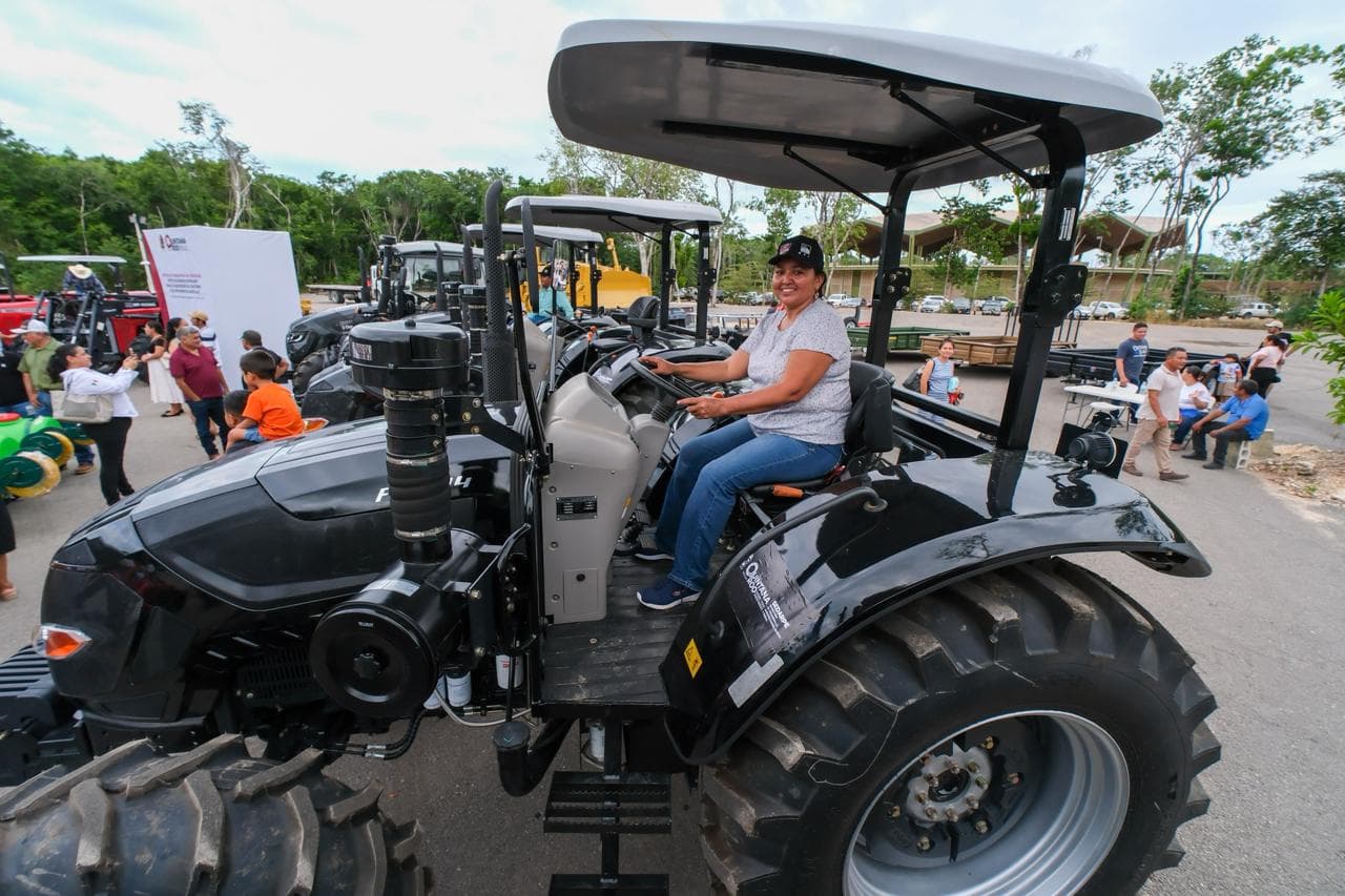 Una mujer operando un tractor moderno de color gris durante un evento de entrega de maquinaria agrícola con asistentes de fondo.