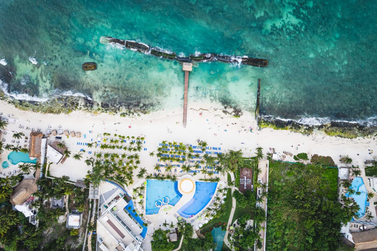 Vista aérea de Playa Petrel Norte en Playa del Carmen mostrando la playa con arena blanca, sombrillas azules, piscinas, palmeras, malecón con estructuras de madera en el agua y turistas en la arena frente al mar turquesa.
