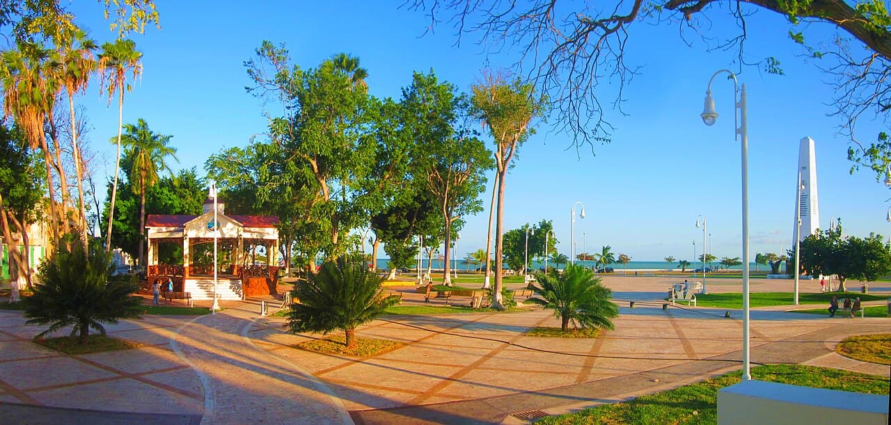 Plaza pública junto al mar en Quintana Roo con áreas verdes, kiosco, farolas y vista al agua bajo cielo despejado.