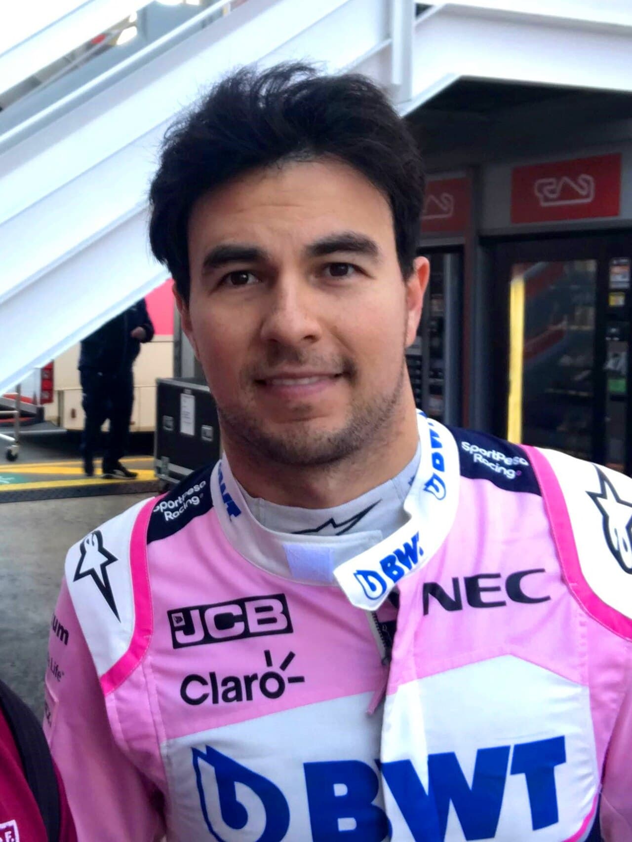 Piloto de automovilismo con uniforme rosa de equipo Cadillac con patrocinadores DBW, NEC, JCB y Claro en paddock de circuito.