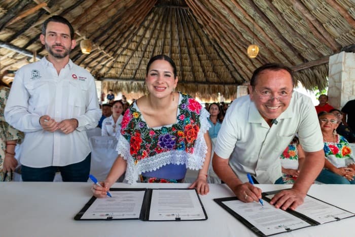 Tres personas firman documentos en una ceremonia bajo una estructura con techo de palma, rodeadas de asistentes en un evento comunitario.