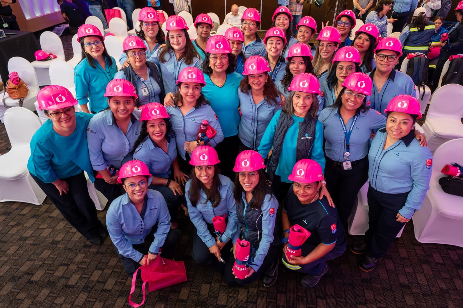 Grupo de mujeres con uniformes azules y cascos de seguridad rosa posando en evento corporativo de Aguakan.