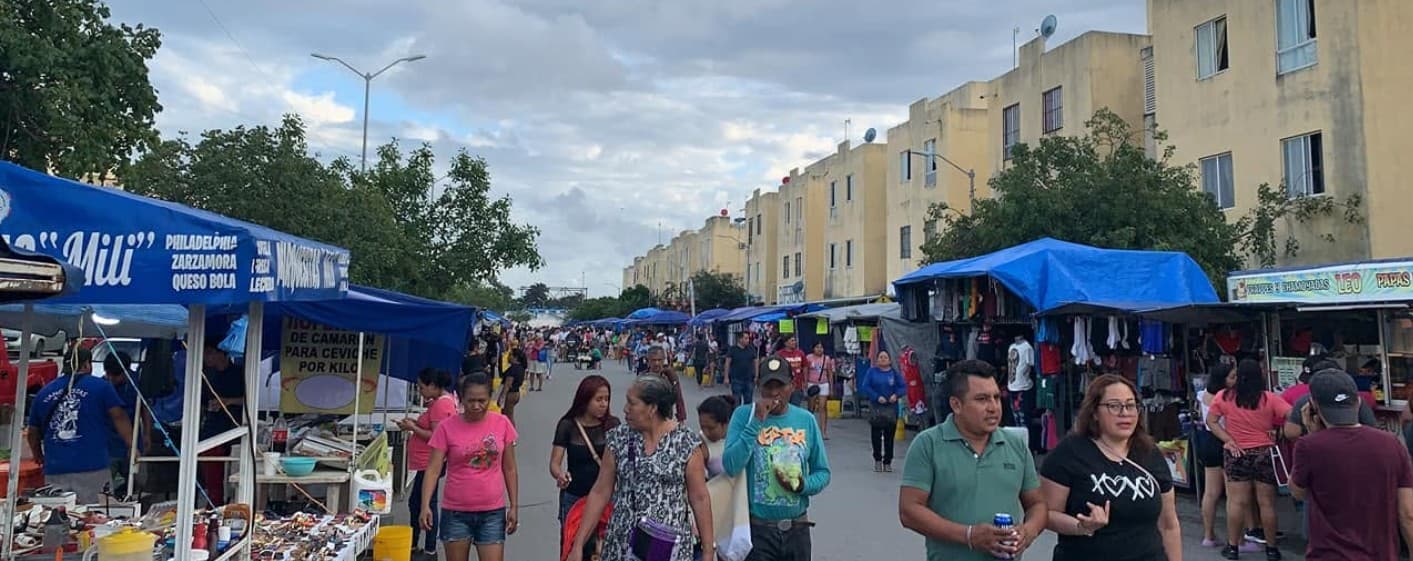 Vista panorámica de un tianguis azul en Cancún con múltiples puestos comerciales, vendedores y público circulando bajo toldos azules, con edificios residenciales de fondo.