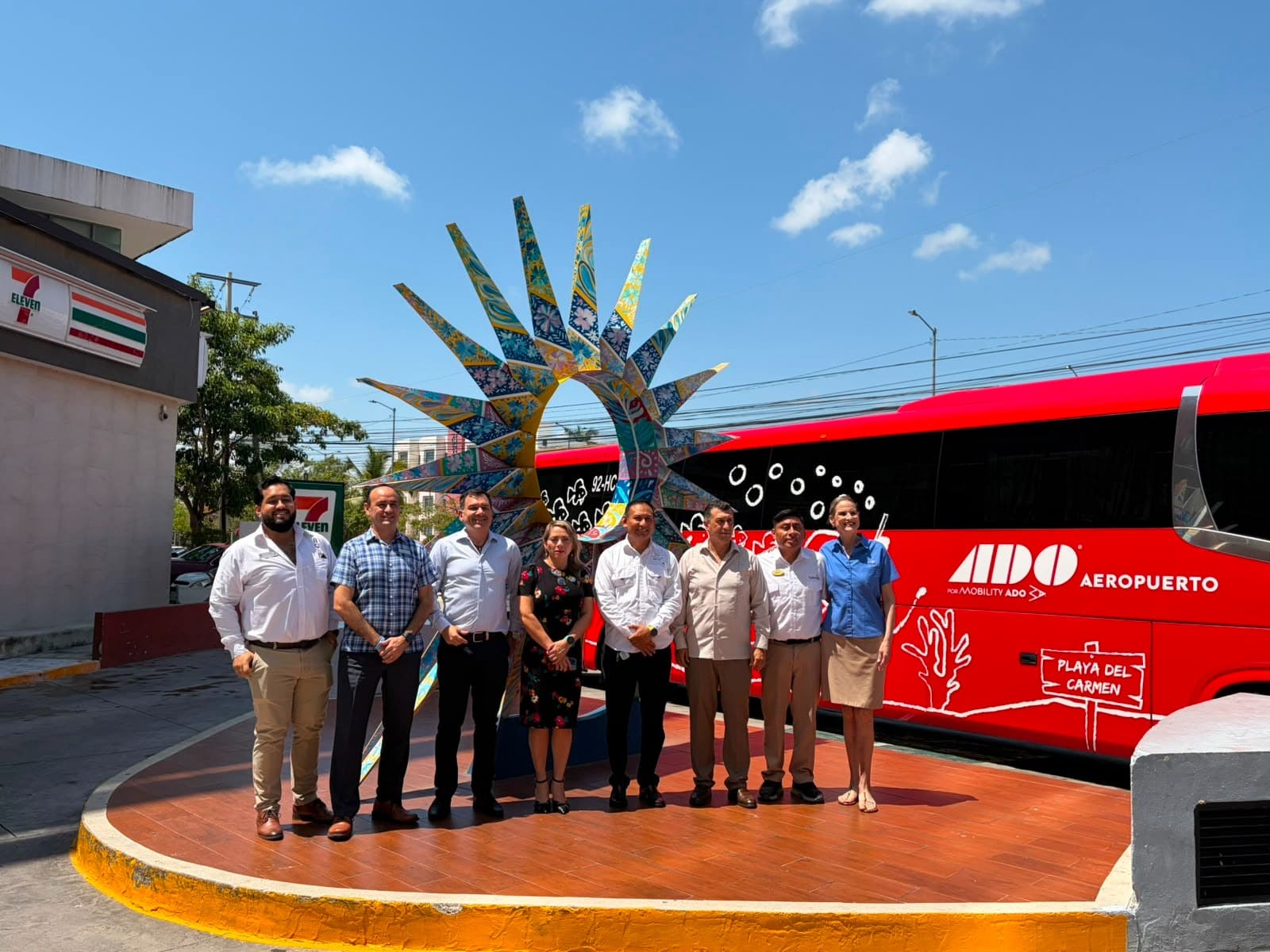 Grupo de personas posando frente a un autobús rojo de ADO Aeropuerto con la escultura de una mano decorada de fondo en Cancún.