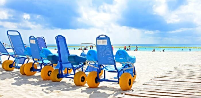 Sillas de ruedas anfibias azules y amarillas estacionadas en la arena de una playa con turistas en el agua al fondo.