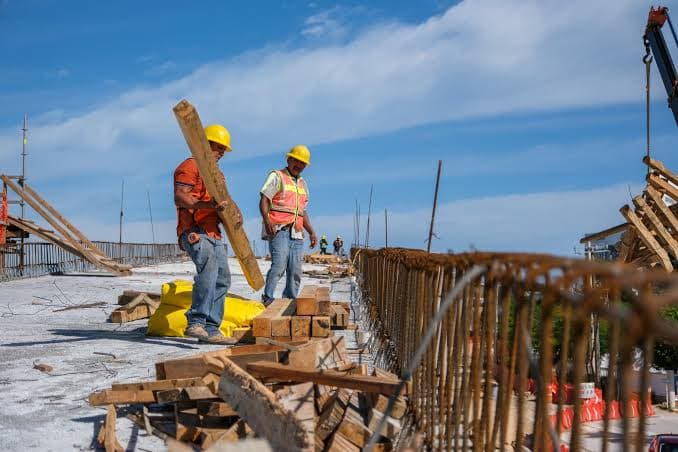 Dos trabajadores de construcción con cascos amarillos y chalecos de seguridad manipulan materiales en una obra en desarrollo.