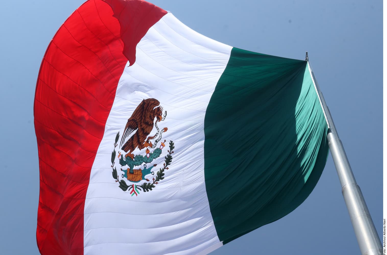 Bandera de México ondeando al viento con el águila y escudo nacional en el centro, contra un cielo azul.