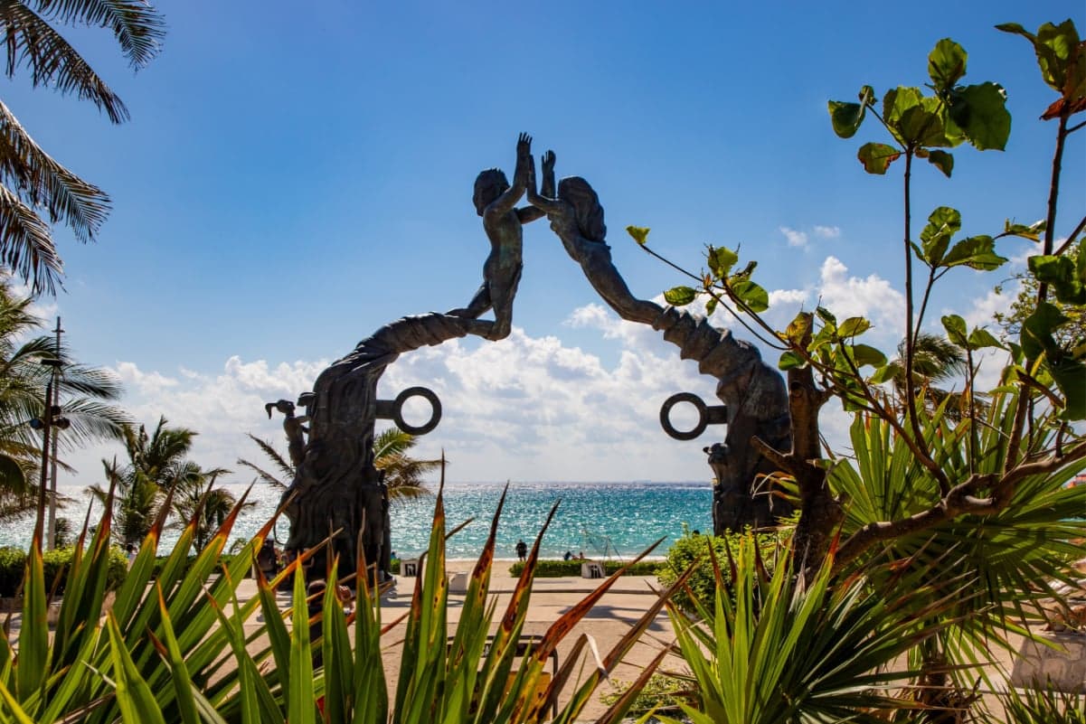 Escultura de dos figuras en la playa de Playa del Carmen con vegetación tropical y mar turqueso de fondo.