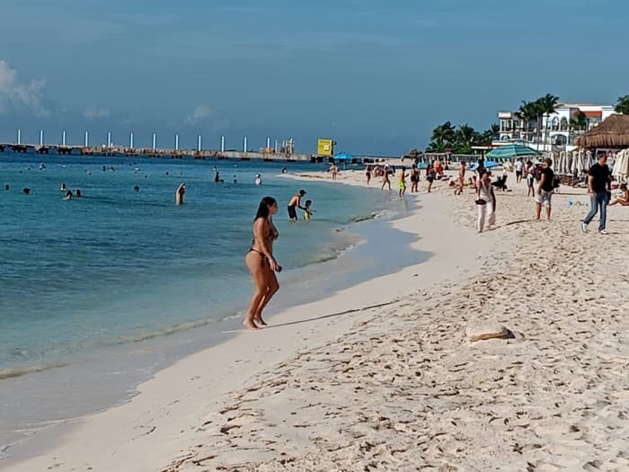 Bañistas y visitantes disfrutan de la playa en Playa del Carmen con aguas turquesas, arena blanca e instalaciones portuarias al fondo.