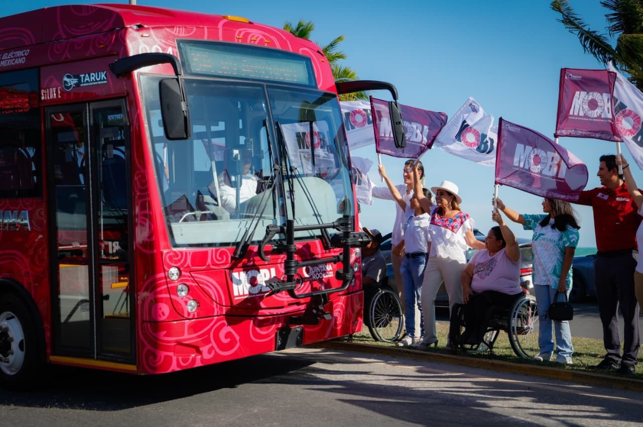 Autobús eléctrico rojo TARUK con decoraciones florales y personas sosteniendo banderas durante un acto de inauguración en Chetumal.