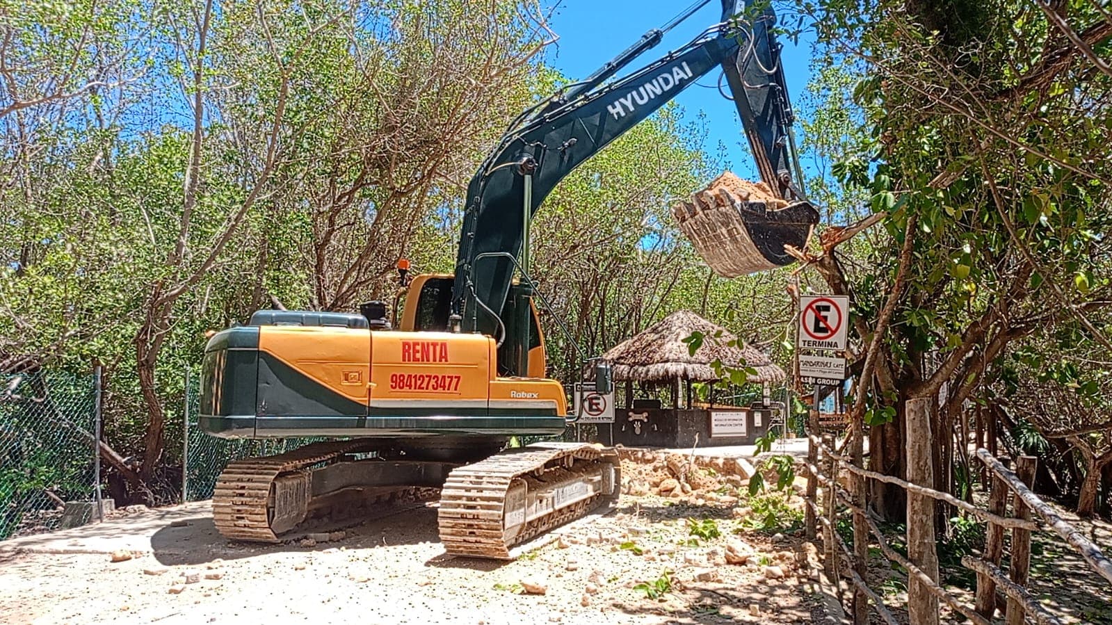 Excavadora Hyundai en operación dentro de un área de playa rodeada de vegetación y árboles, con señalización de no estacionamiento visible.