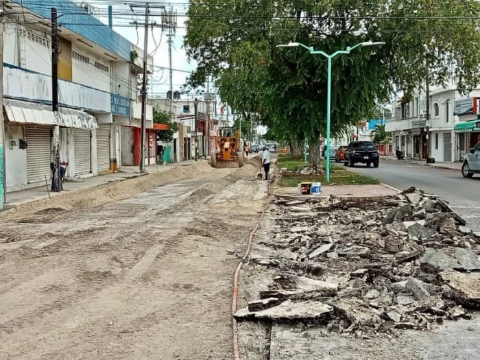 Calle en construcción o reparación con escombros y materiales de obra, edificios comerciales a los lados y señalética urbana.