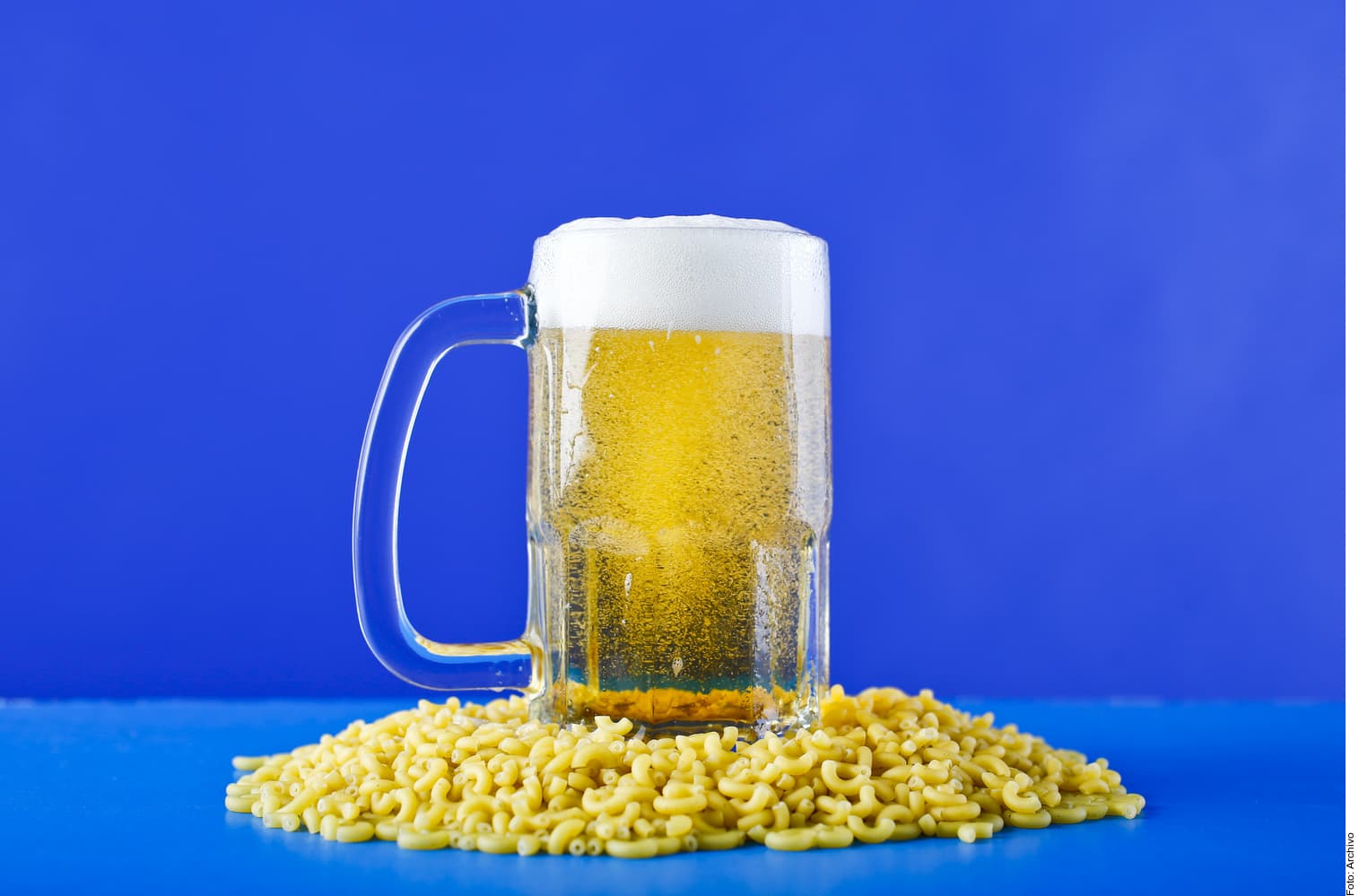 Jarra de cerveza con espuma junto a un montículo de cereal tipo aro sobre fondo azul.