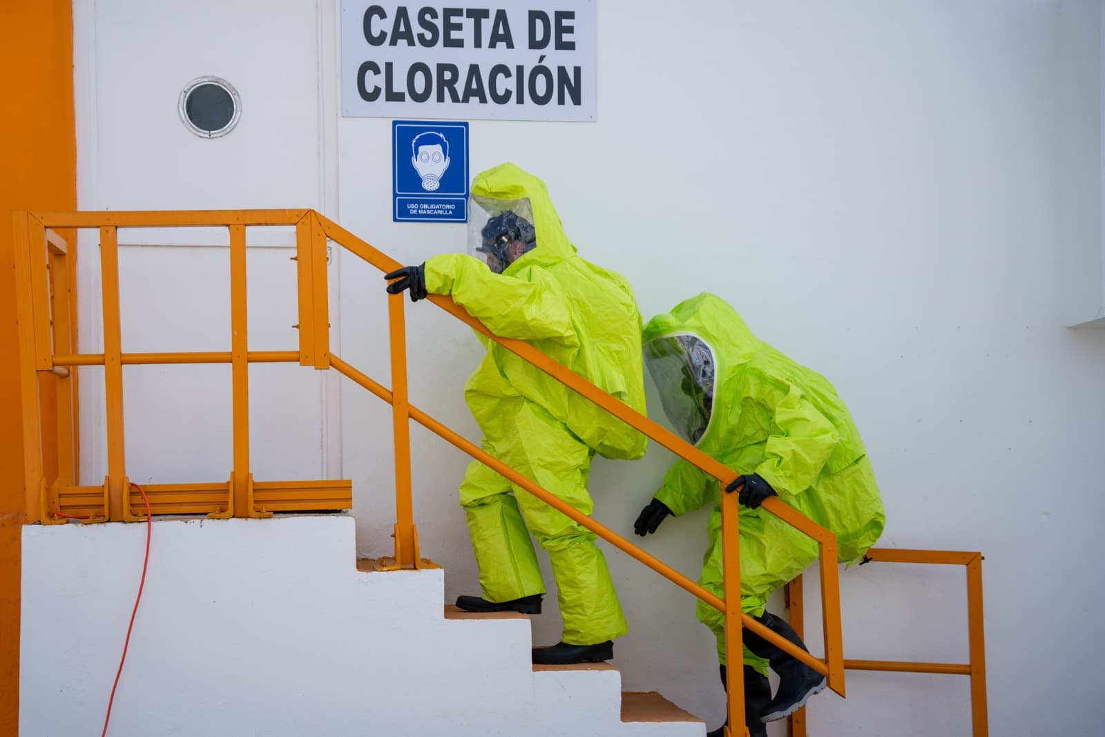 Dos operarios con trajes de protección amarillo fluorescente descienden escaleras metálicas junto a una caseta de cloración durante un simulacro de emergencia.