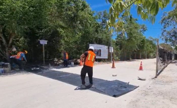 Trabajador en chaleco naranja instala una malla o tela en la arena de una playa, con árboles de fondo y señalización de obra visible.
