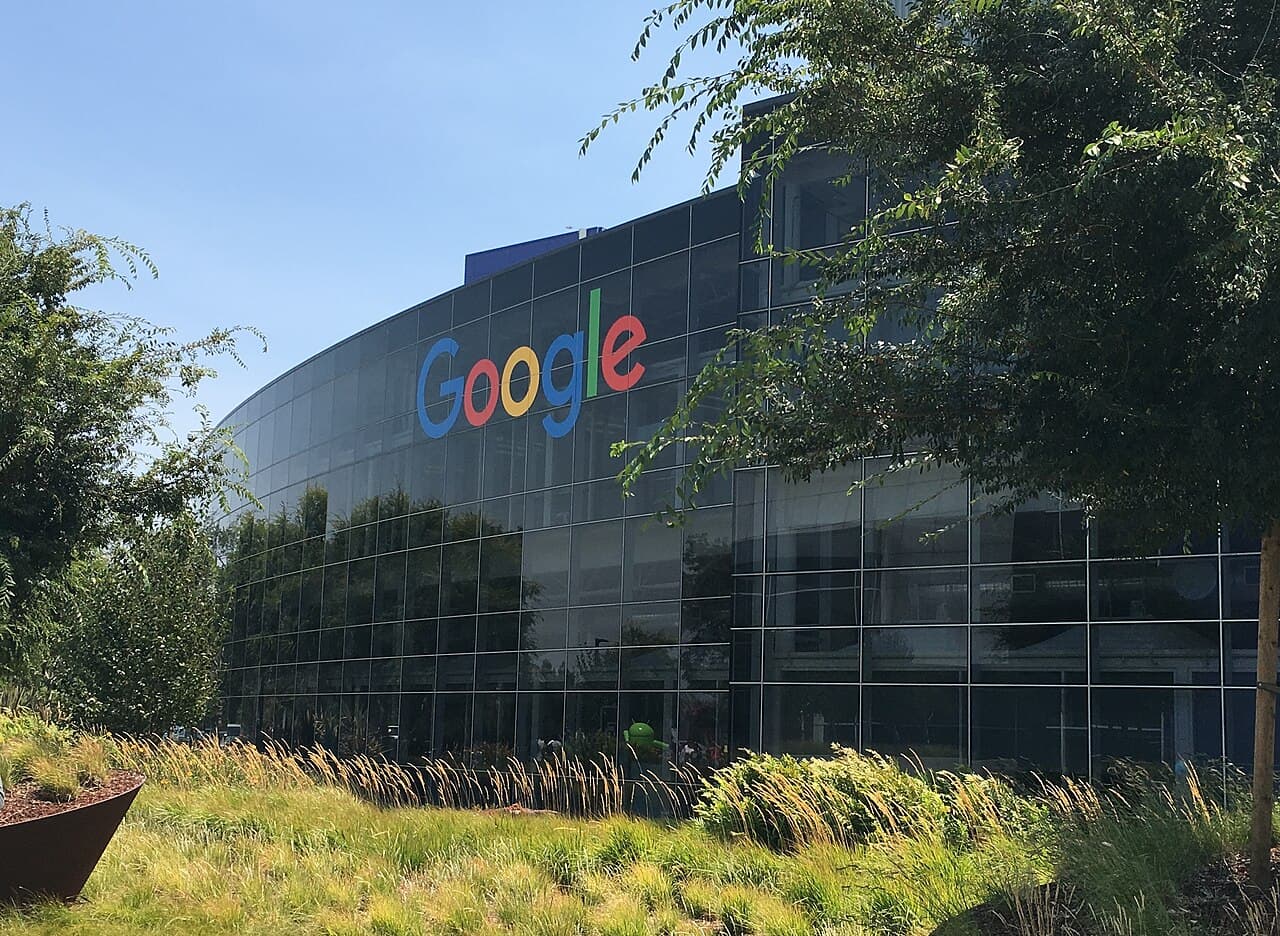 Edificio de oficinas con el logo de Google en su fachada de vidrio, rodeado de vegetación.