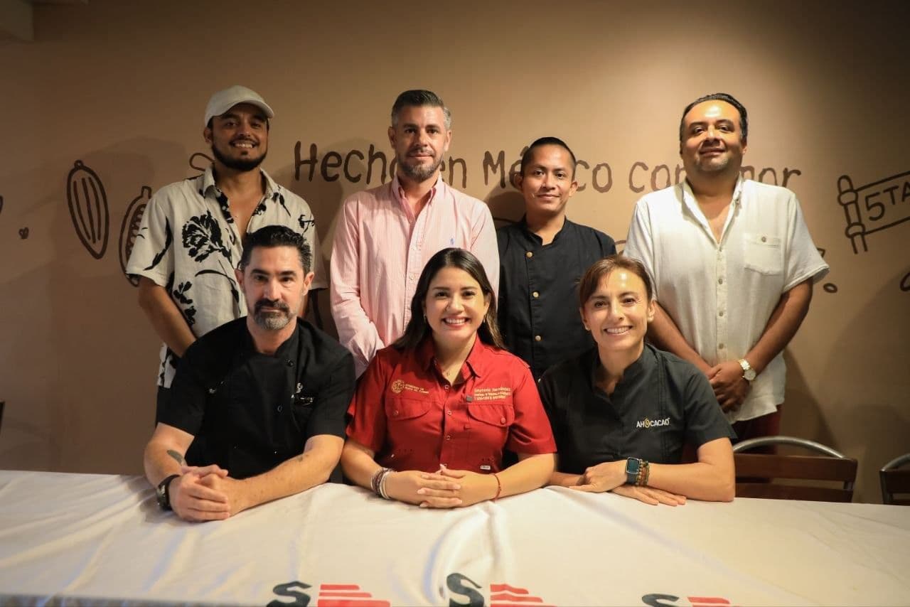 Grupo de ocho personas posando juntas frente a una pared con texto sobre Playa del Carmen, varios vistiendo ropa formal y de colores variados.