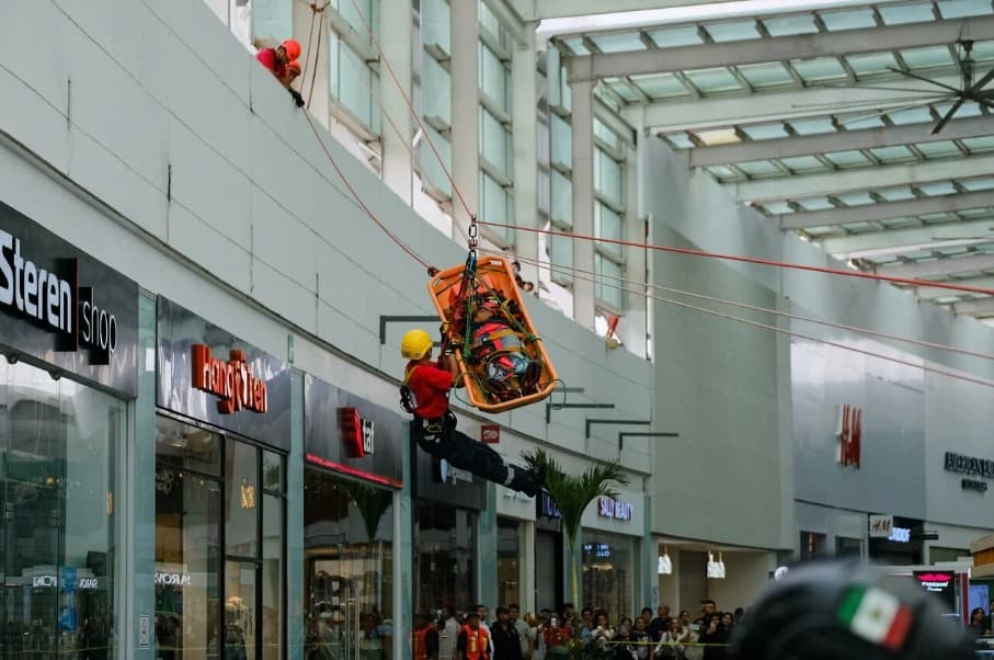 Rescatista en arnés desciende en camilla de evacuación dentro de un centro comercial durante un simulacro de emergencia.