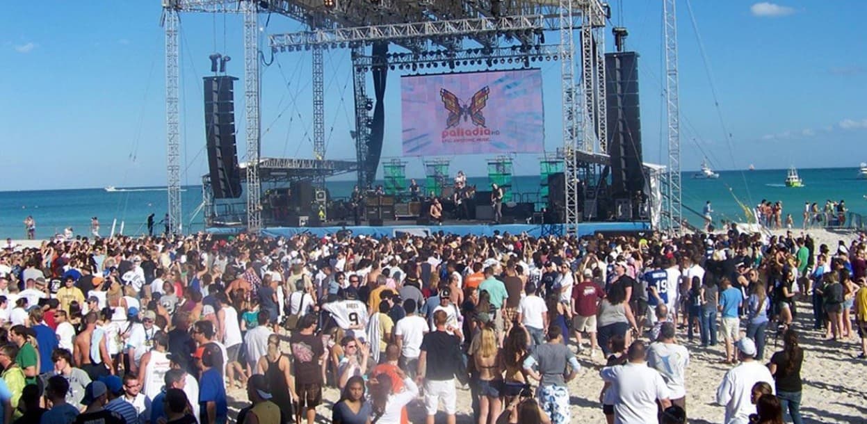 Multitud de turistas en playa durante evento musical con escenario y pantalla gigante frente al mar en la Riviera Maya.
