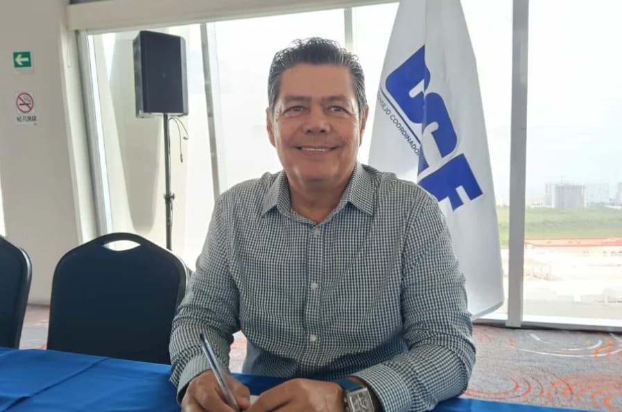 Hombre de mediana edad en camisa cuadriculada sonriendo durante una conferencia o rueda de prensa con bandera azul de fondo.