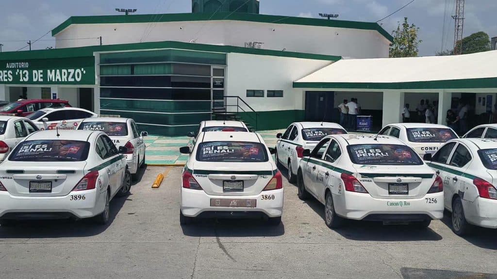 Flota de taxis blancos con distintivos de la empresa estacionados frente a un auditorio con franjas verdes en Cancún.
