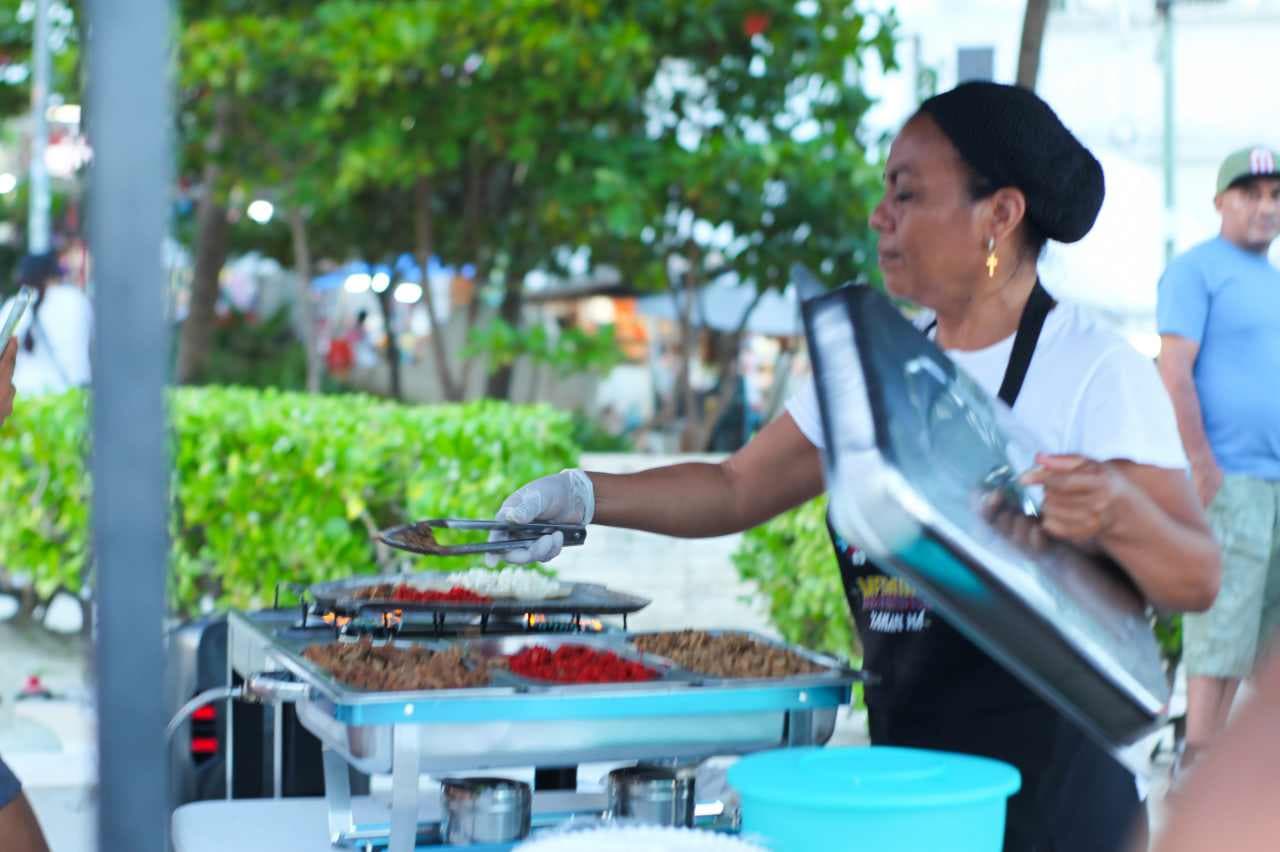 Emprendedora sirve alimentos en un evento gastronómico callejero bajo árboles en ambiente de parque público.