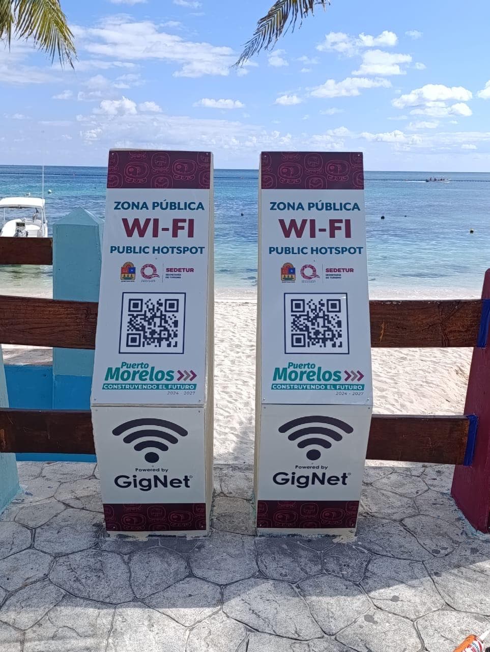 Dos señalamientos de zona pública de Wi-Fi en una playa del Caribe con el mar y cielo azul de fondo.