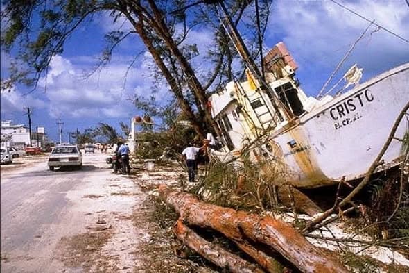 Barco de pesca volcado sobre tierra con árbol derribado y edificios dañados de fondo, resultado del paso de un huracán en zona costera.