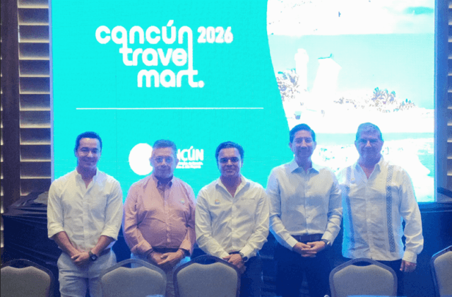 Cinco hombres de traje blanco posados frente a pantalla con logo de Cancún Travel Mart 2024.