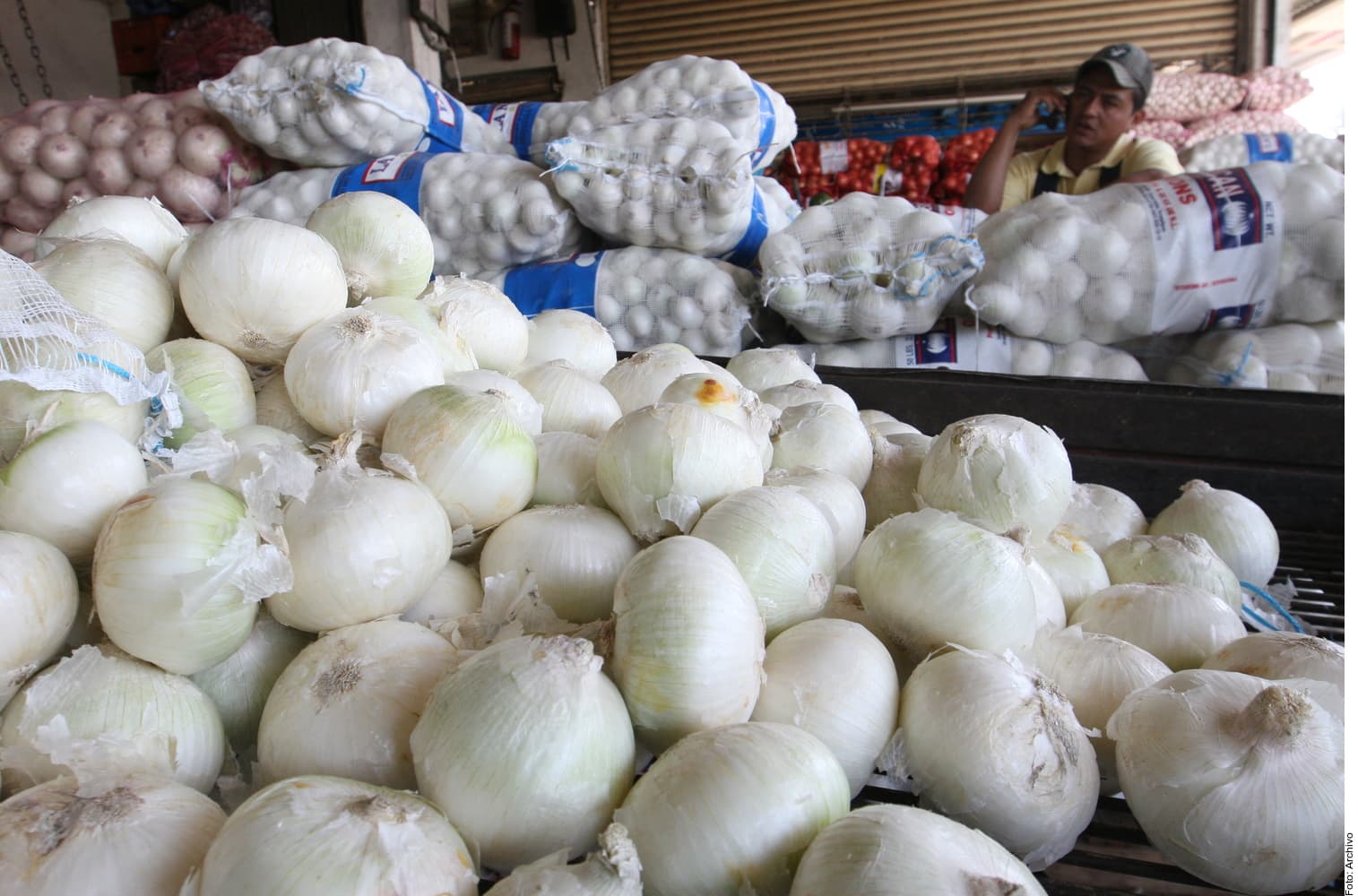 Cebollas blancas frescas exhibidas en un puesto de mercado con bolsas de ajo al fondo y un vendedor atendiendo.
