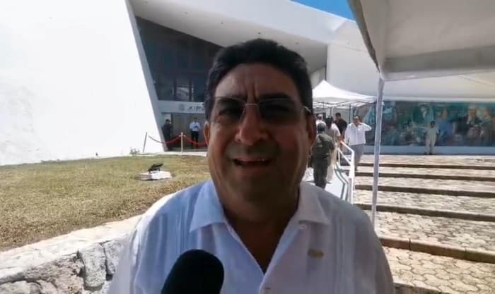 Manuel Moguel Manzur sonríe ante un micrófono en un evento al aire libre con carpas y asistentes de fondo.