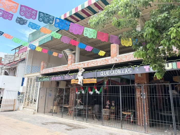 Restaurante con letrero de Quesadillas y decoraciones coloridas en fachada de ladrillo, con local cerrado al público mediante rejas metálicas.