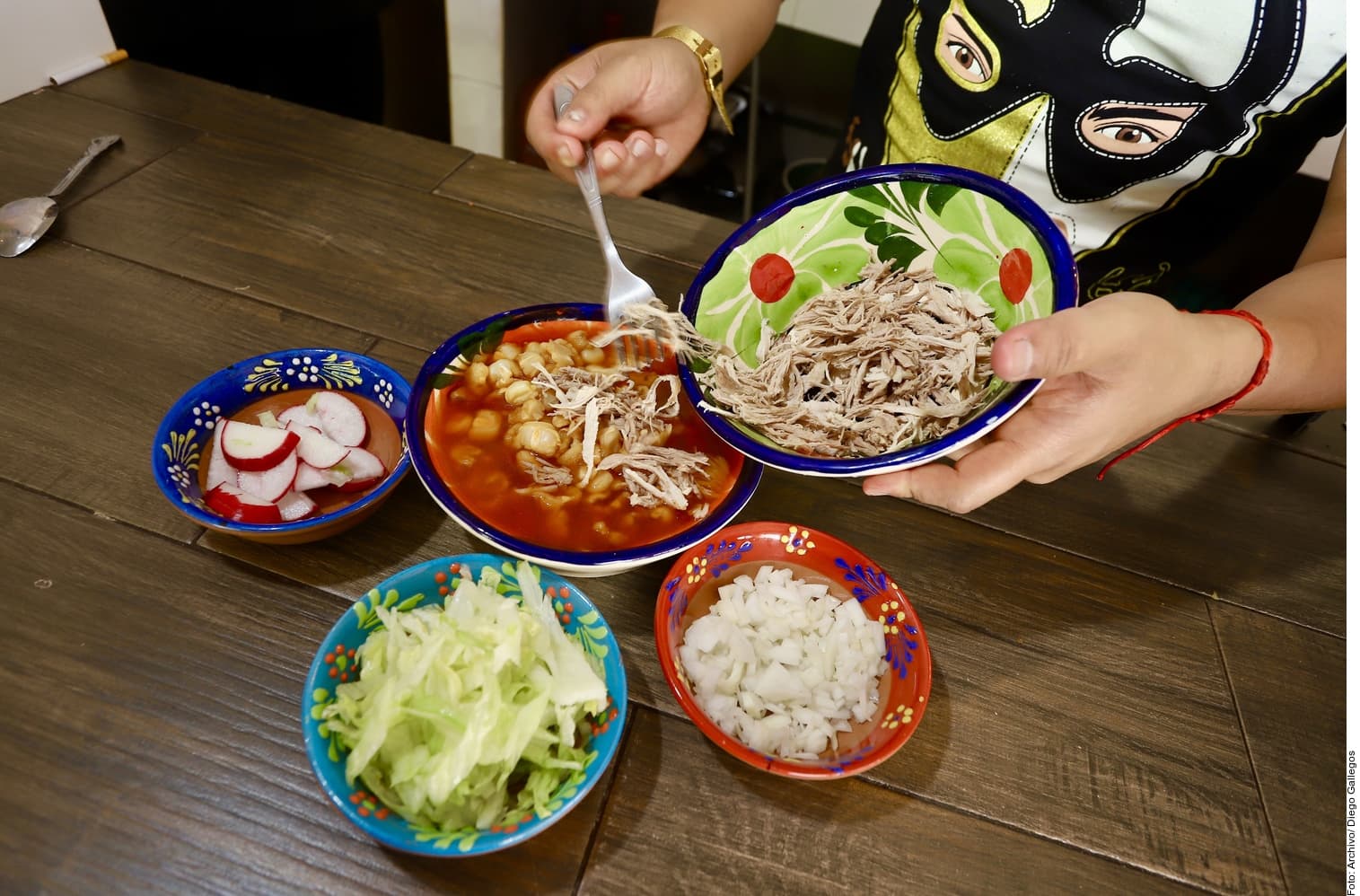 Persona sosteniendo platos con ingredientes frescos como carne desmenuzada, verduras, cebolla y otros componentes de un plato tradicional mexicano sobre una mesa de madera.