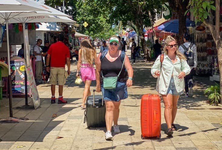 Turistas con maletas caminan por una calle comercial del centro de Cancún bajo sombrillas y árboles, con tiendas y vendedores ambulantes a los lados.