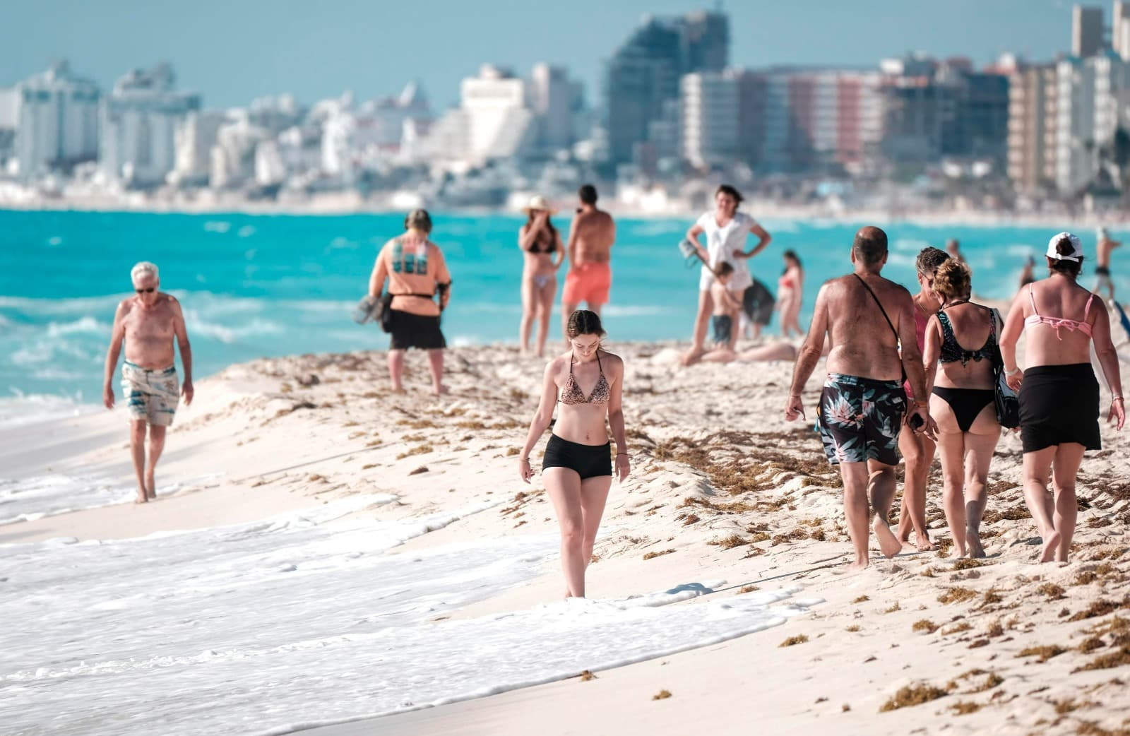 Bañistas en una playa turística con aguas turquesas y edificios de ciudad al fondo.